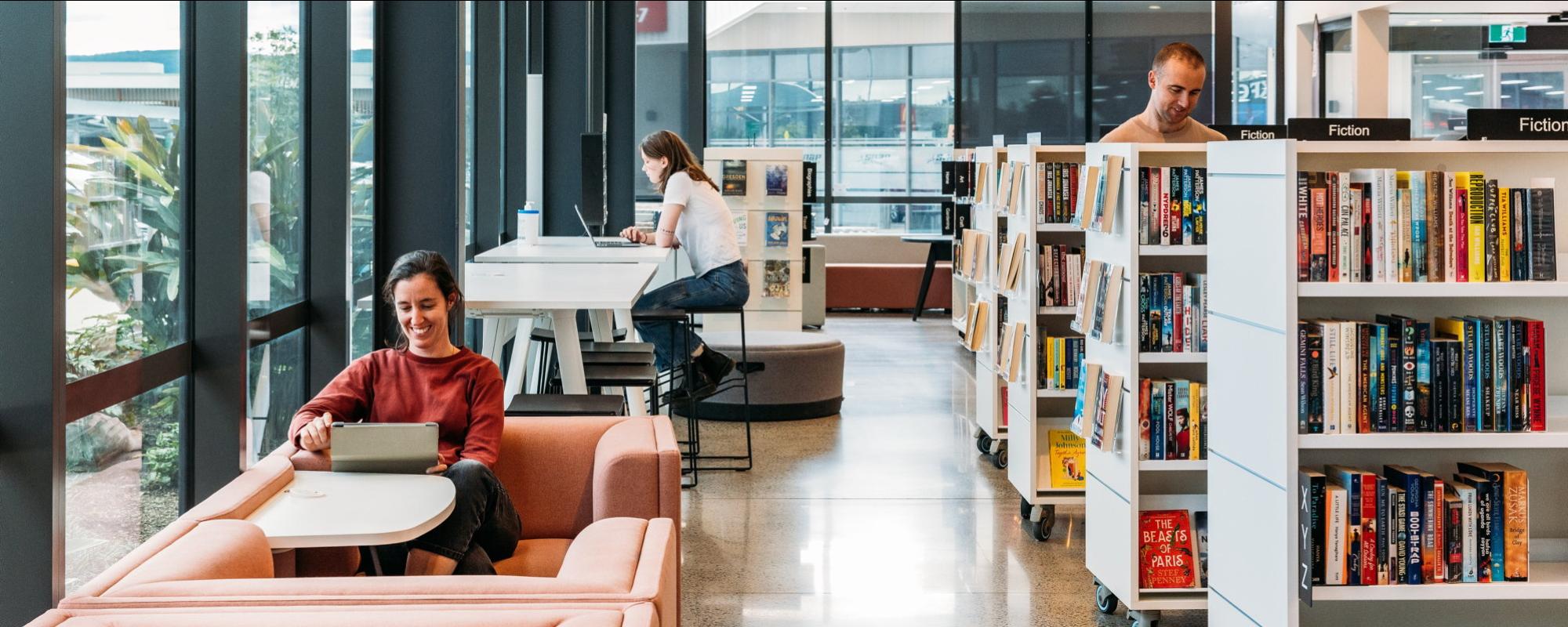 people sitting in a library using devices and looking at book shelves